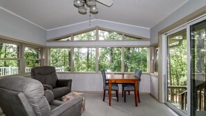 Ein Wintergarten mit Deckenventilator, einem Esstisch mit Stühlen, einem grauen Sofa und großen Fenstern mit Blick auf Bäume.