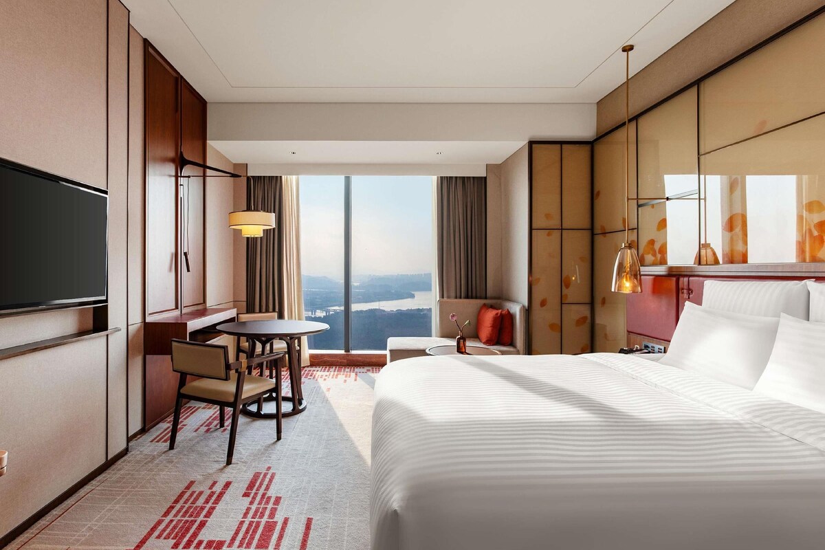 A hotel room with a bed, a desk, a chair, a TV, and a large window offering a view.