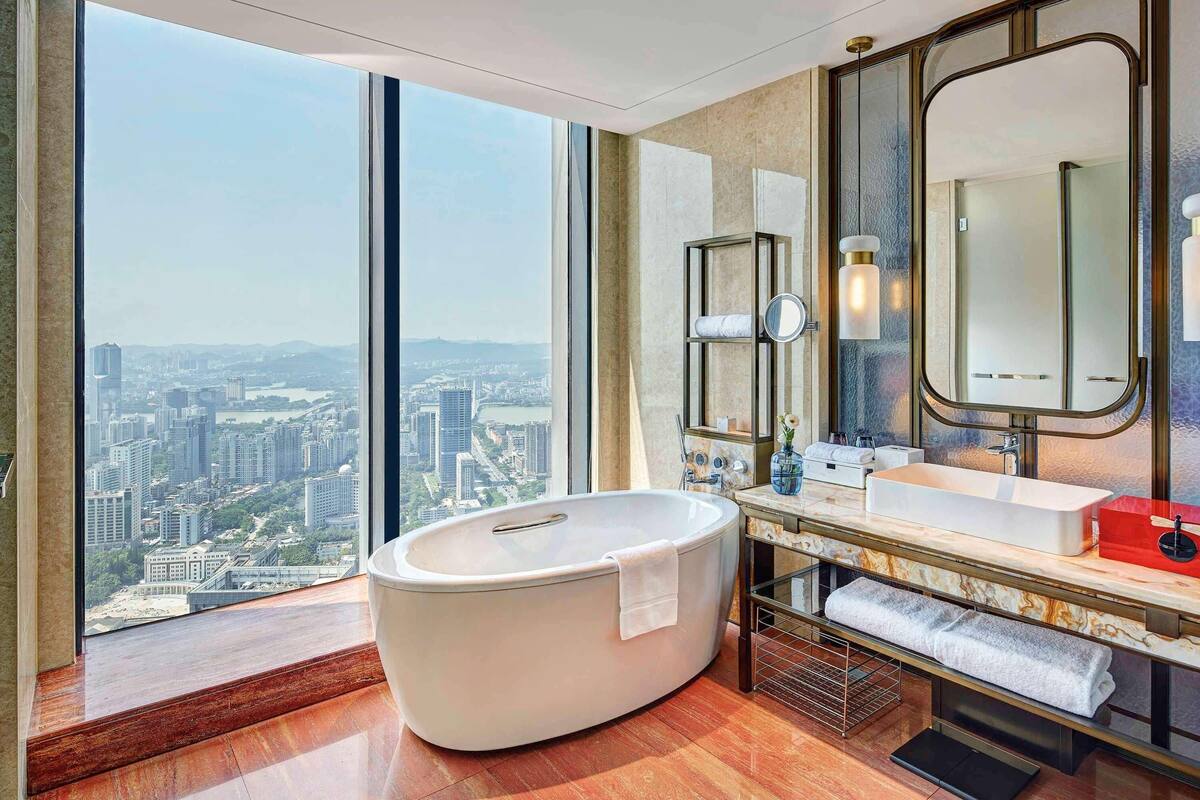Casa de banho moderna com uma banheira grande, toucador com espelho e vista para a paisagem urbana através de grandes janelas.