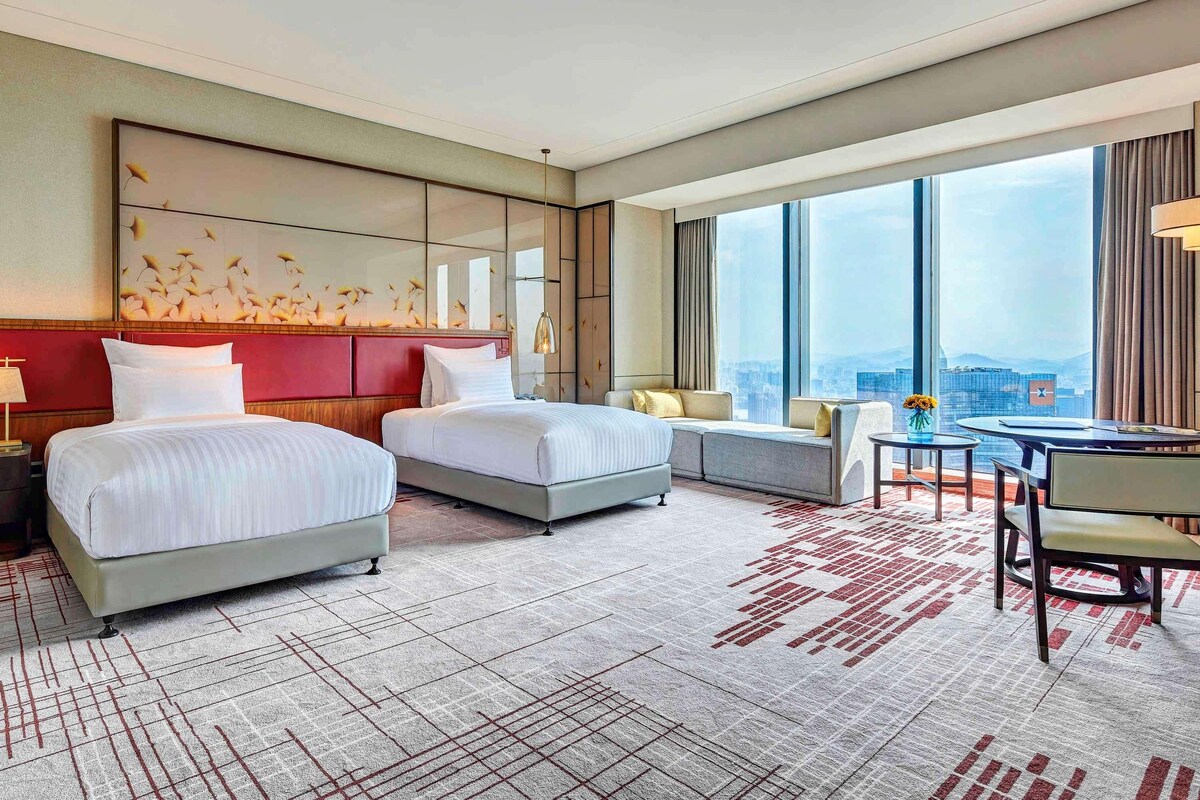 A hotel room with two beds, a sofa, a dining area, and a large window offering a city view.