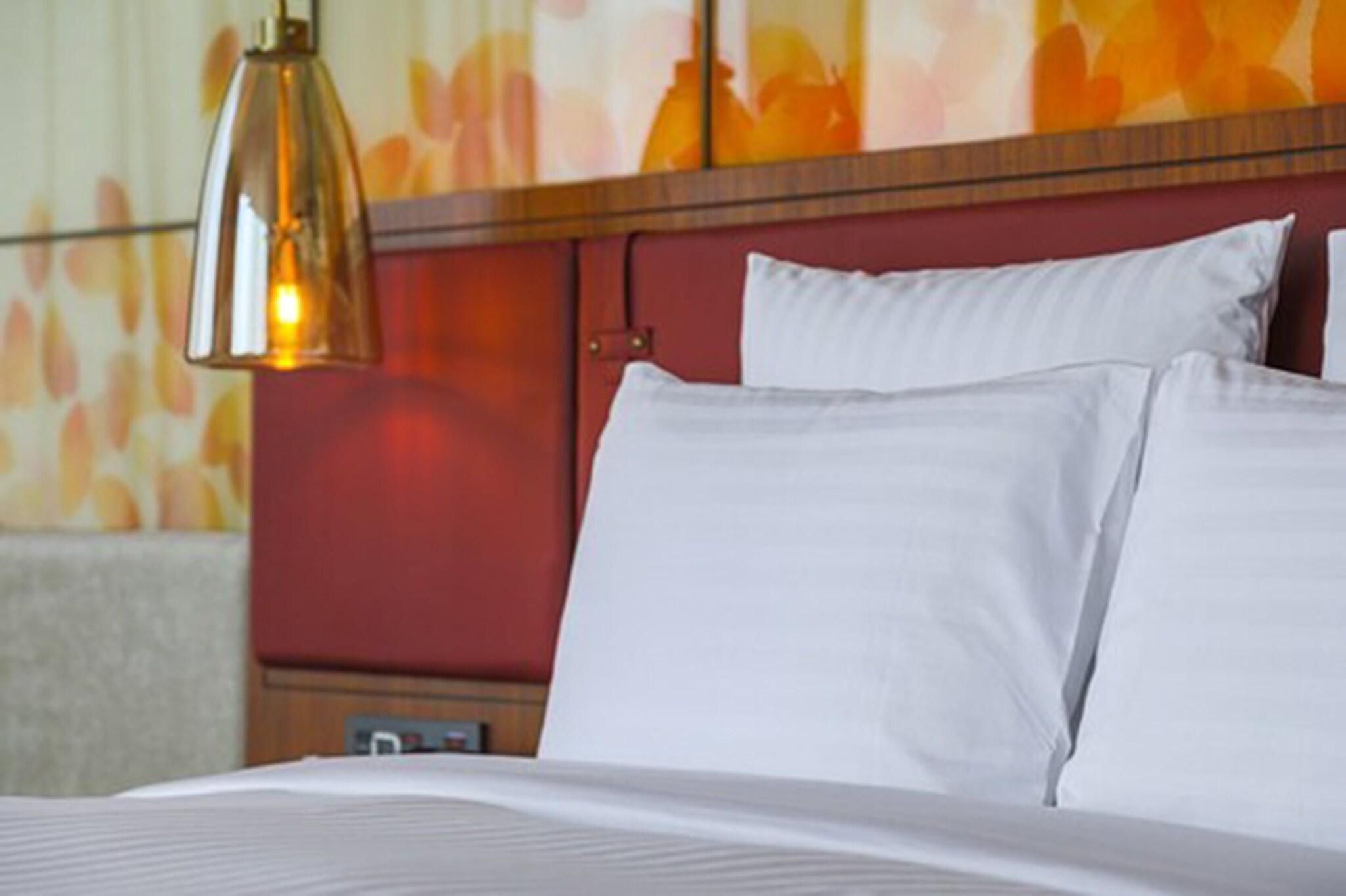A hotel room with a bed, white pillows, a wooden headboard, and a hanging pendant light.