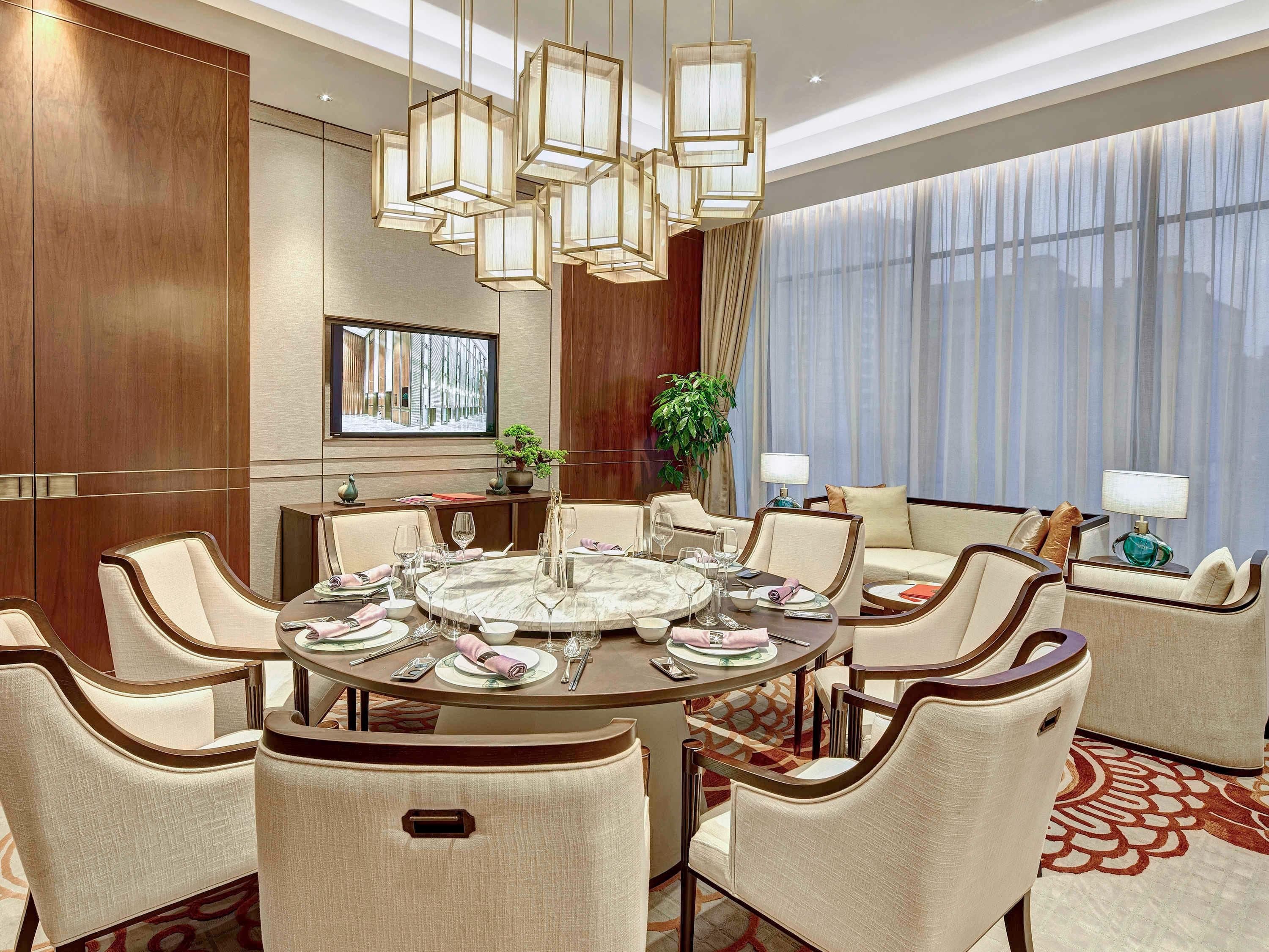A hotel dining room with a round table, multiple chairs, a sofa, and a large window with curtains.