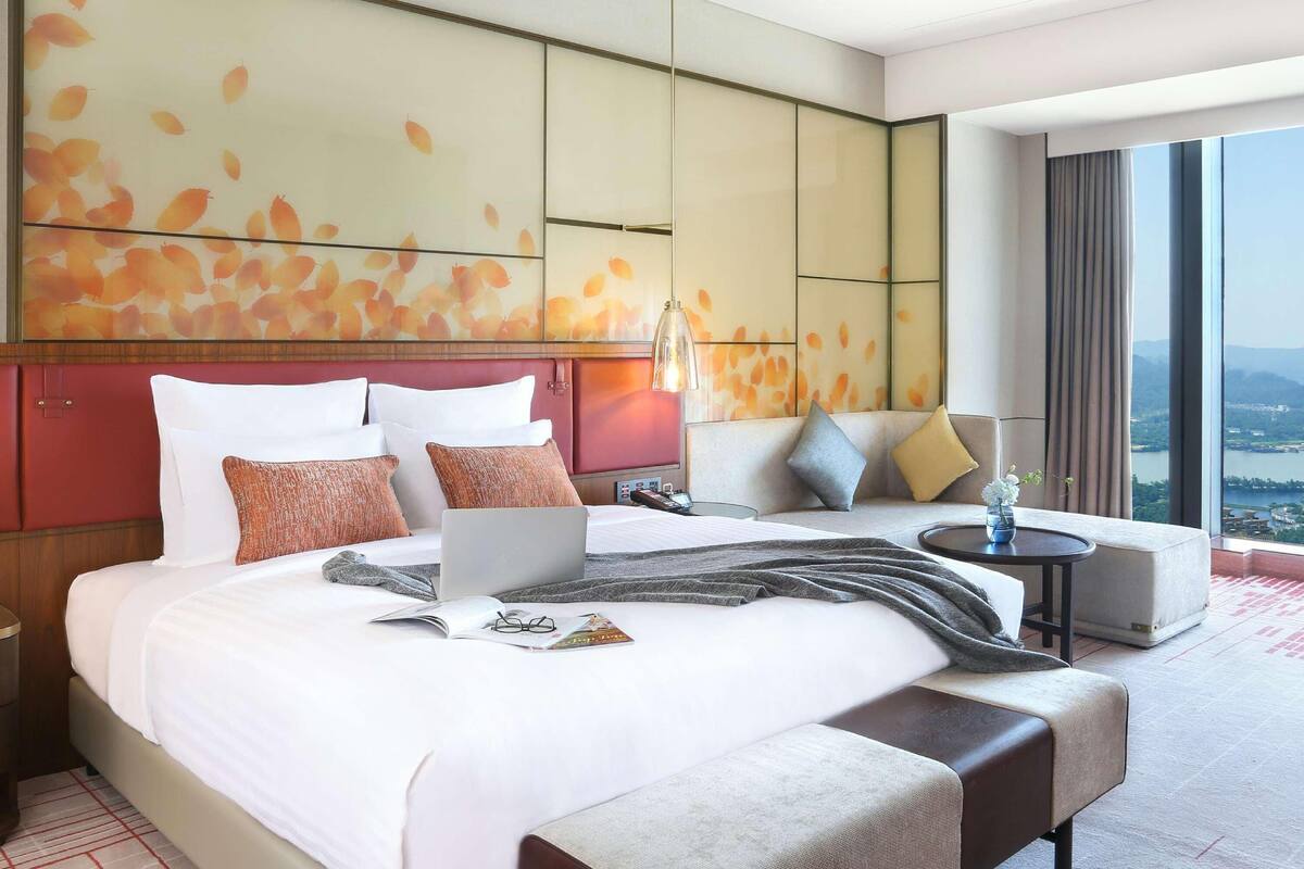 Quarto de hotel moderno com uma cama grande, área de estar e vista para um lago.