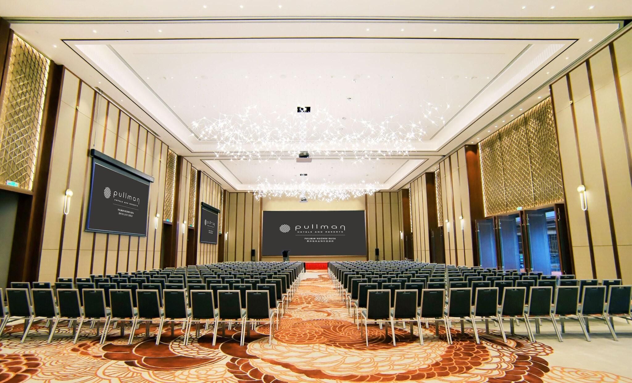 Un salón de conferencias con filas de sillas, un escenario y una pantalla grande que muestra el nombre del hotel.