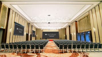 Un salón de conferencias con filas de sillas, un escenario y una pantalla grande que muestra el nombre del hotel.