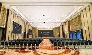 Une salle de conférence avec des rangées de chaises, une scène et un grand écran affichant le nom de l’hôtel.