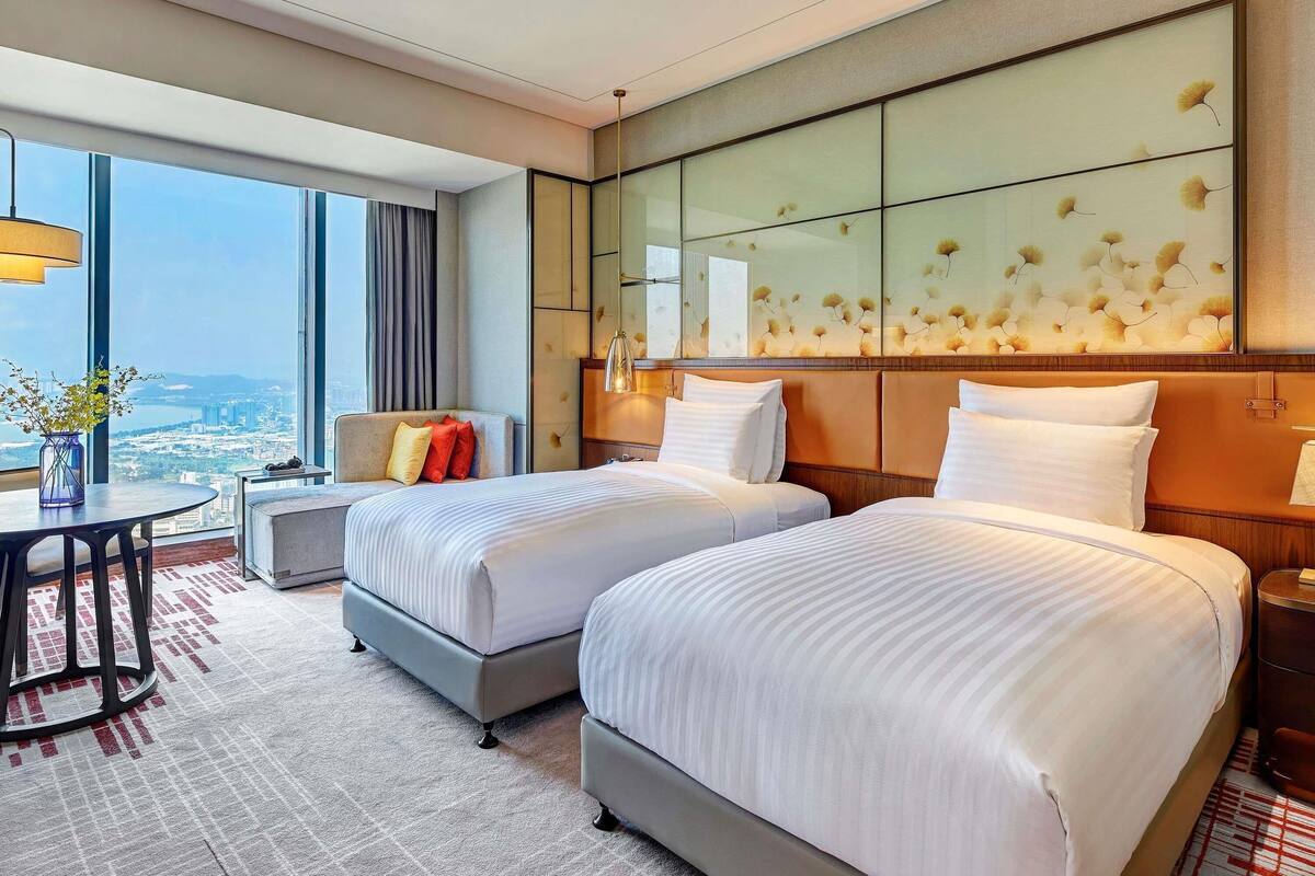 Quarto de hotel com duas camas, uma mesa de jantar, um sofá e uma grande janela com vista para a cidade.