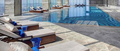 A modern indoor pool area with sun loungers, a clear pool, and large windows.