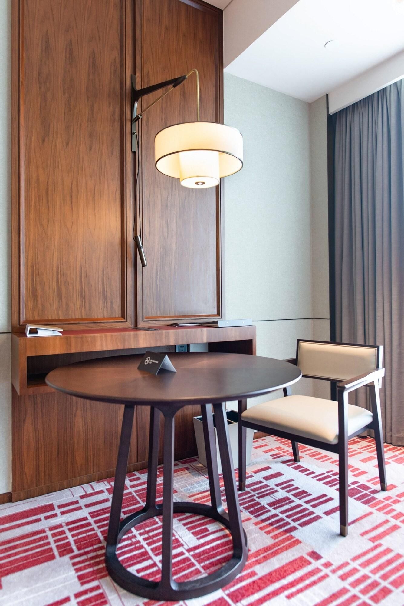 A hotel room with a wooden cabinet, a round table with a chair, a hanging lamp, and a patterned carpet.