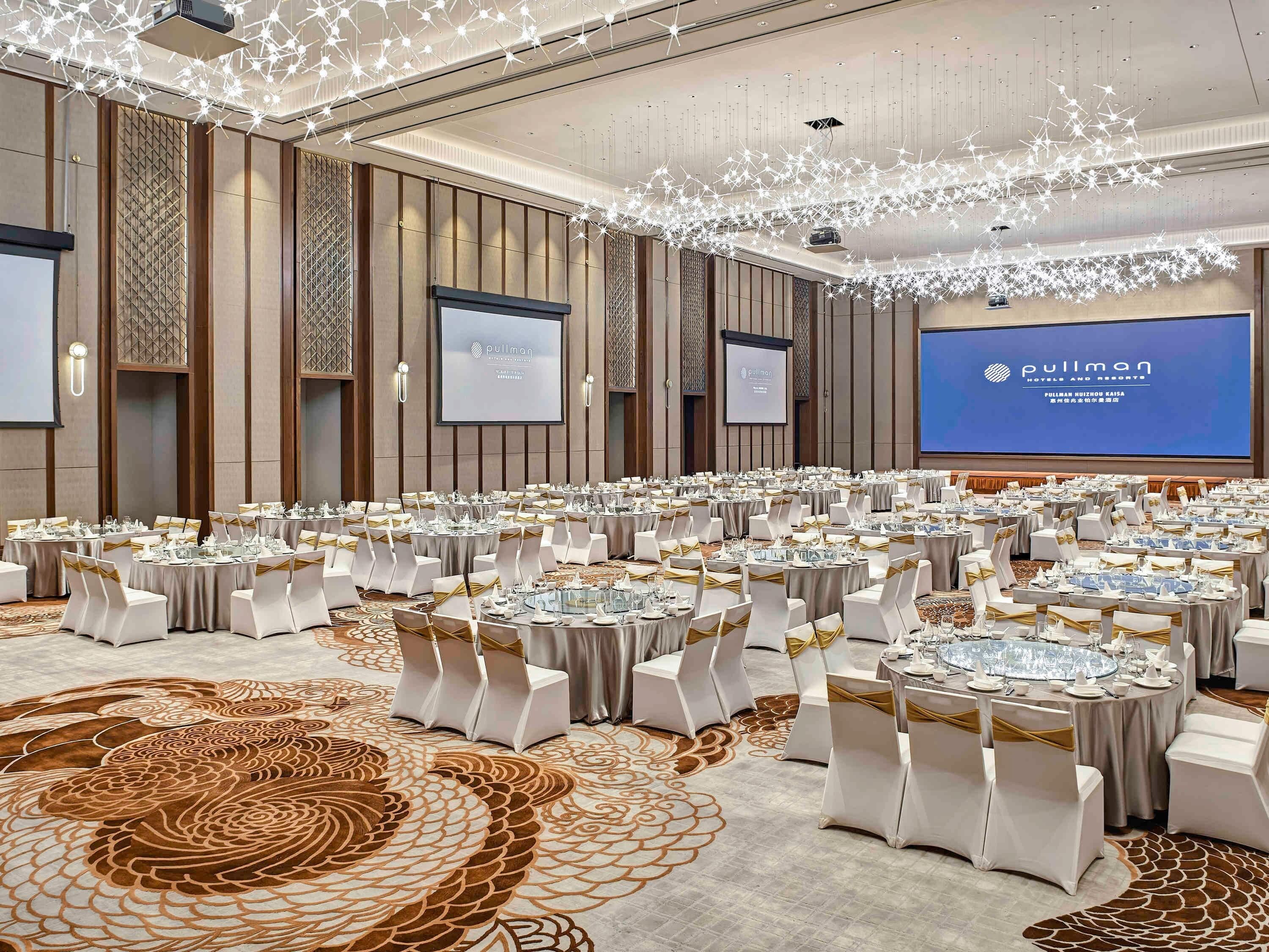 Un amplio salón de conferencias con mesas redondas, sillas blancas y una pantalla grande que muestra el nombre del hotel.
