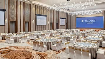 Un amplio salón de conferencias con mesas redondas, sillas blancas y una pantalla grande que muestra el nombre del hotel.
