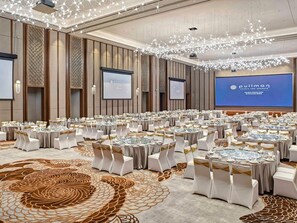 Une grande salle de conférence équipée de tables rondes, de chaises blanches et d’un grand écran affichant le nom de l’hôtel.