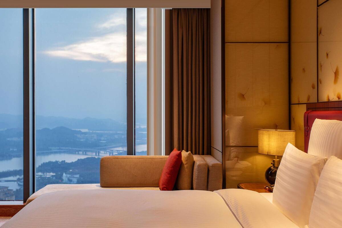 Quarto de hotel com uma janela grande, uma cama, uma mesa de cabeceira e uma luminária.