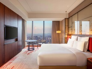 A hotel room with a large bed, a TV mounted on the wall, a small table and chairs, and a city view through large windows.