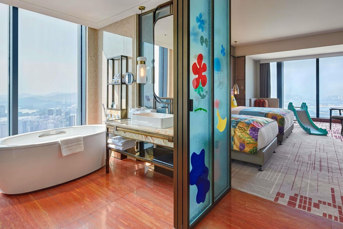 A modern hotel room with a large bathtub, a separate bathroom, a bed with a colorful bedspread, and a large window offering a city view.