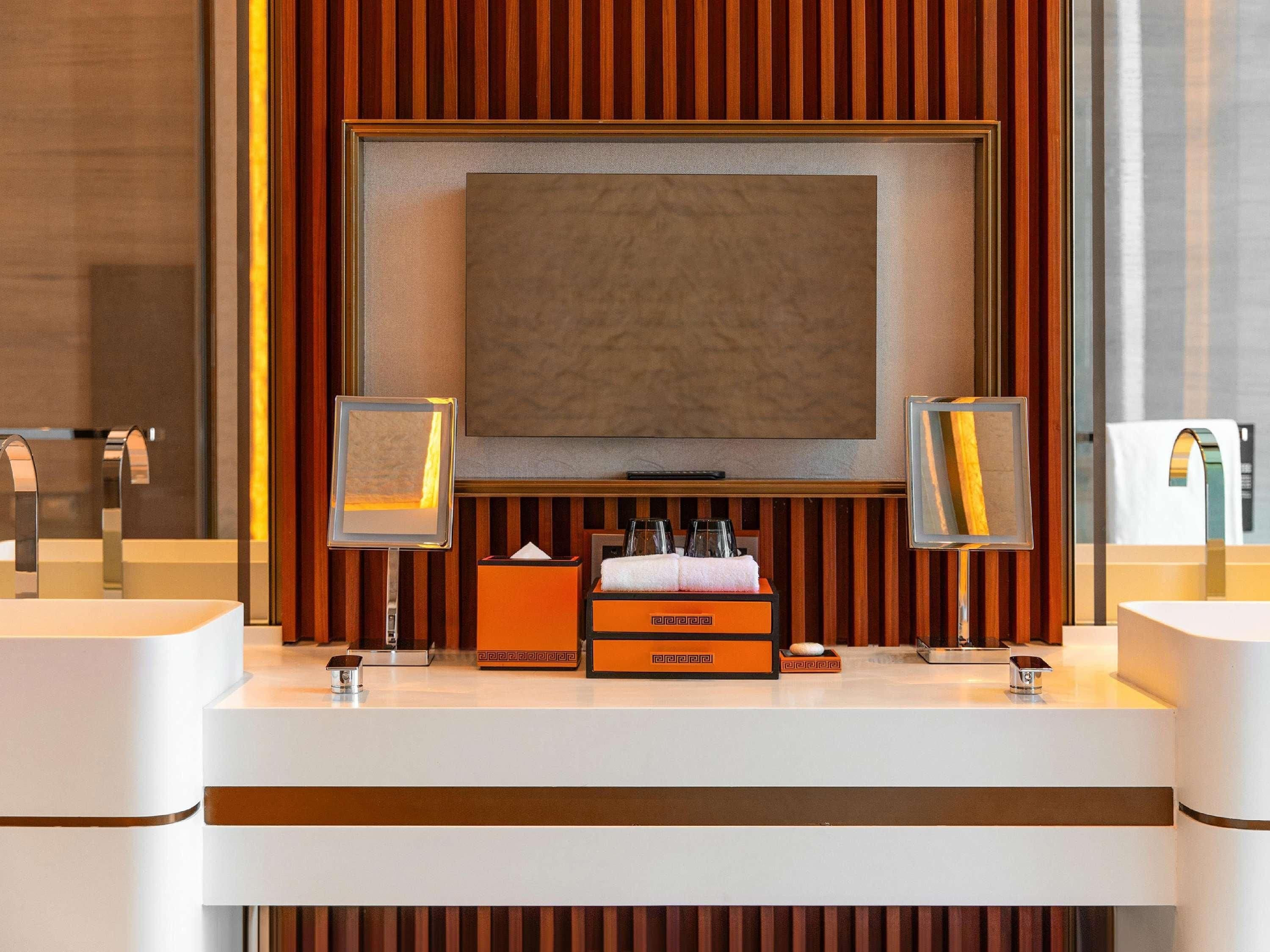 A modern bathroom with a large mirror, two sinks, a vanity with toiletries, and a television above the mirror.