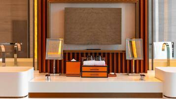 A modern bathroom with a large mirror, two sinks, a vanity with toiletries, and a television above the mirror.