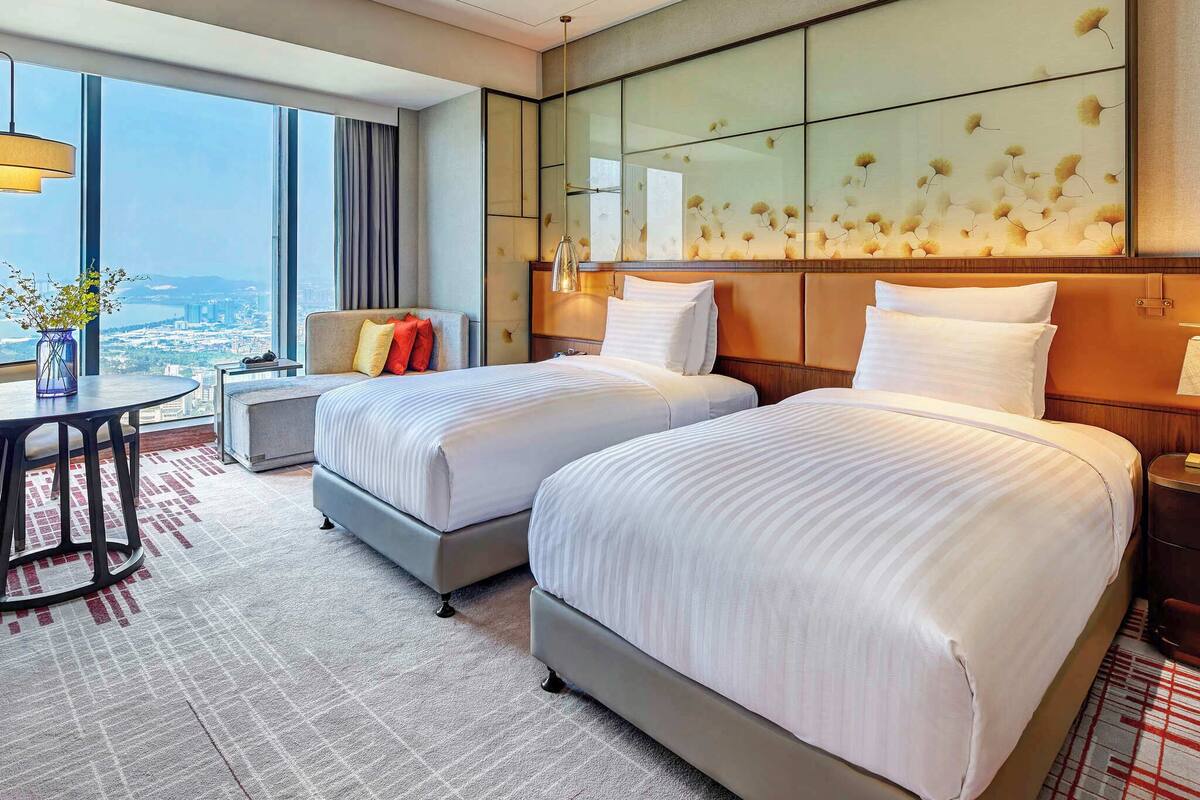 Quarto de hotel com duas camas, uma escrivaninha, uma cadeira, uma janela grande com vista para a cidade e um painel decorativo na parede.