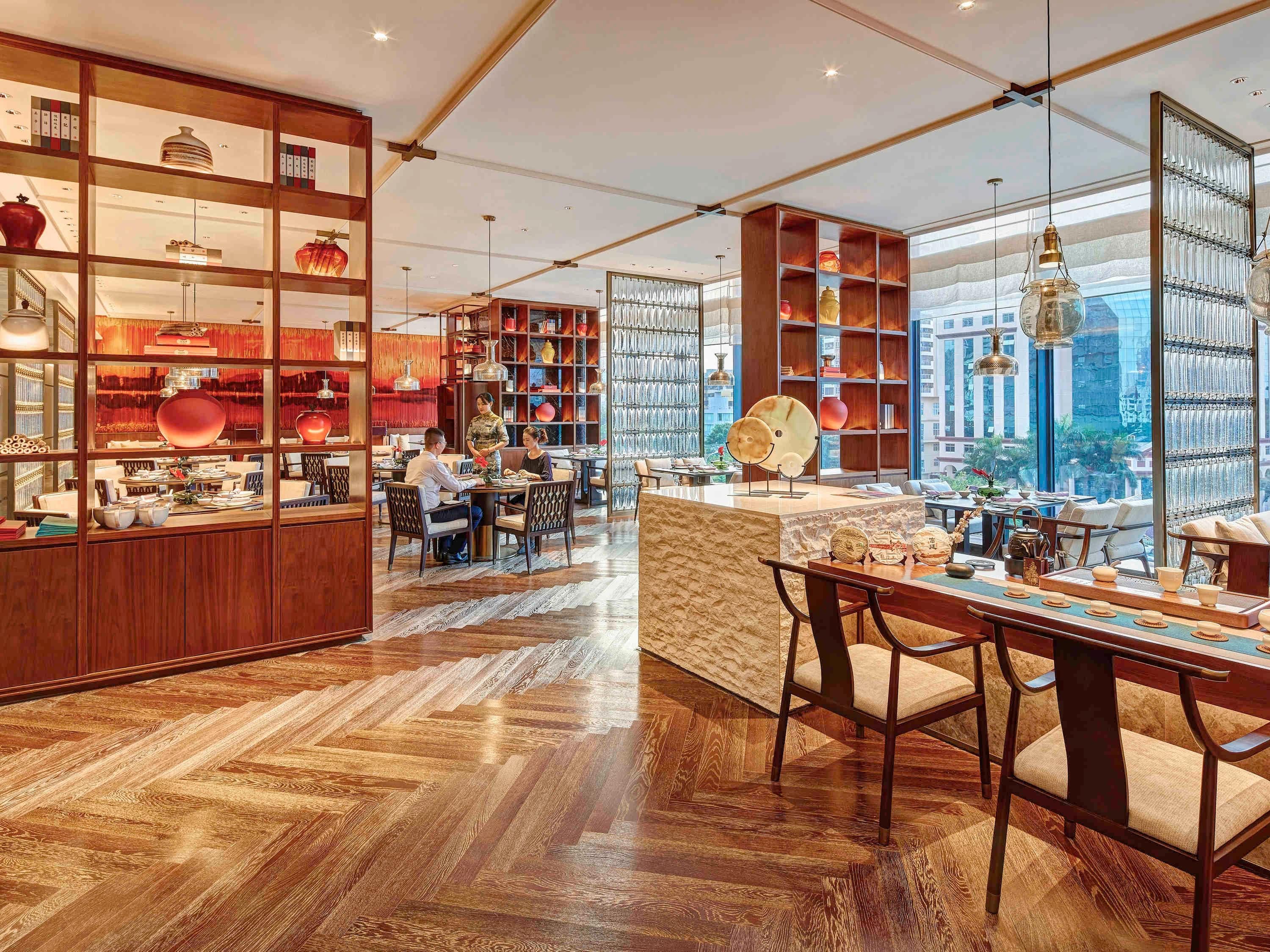 A spacious hotel lobby with wooden flooring, a central seating area, and large windows offering city views.