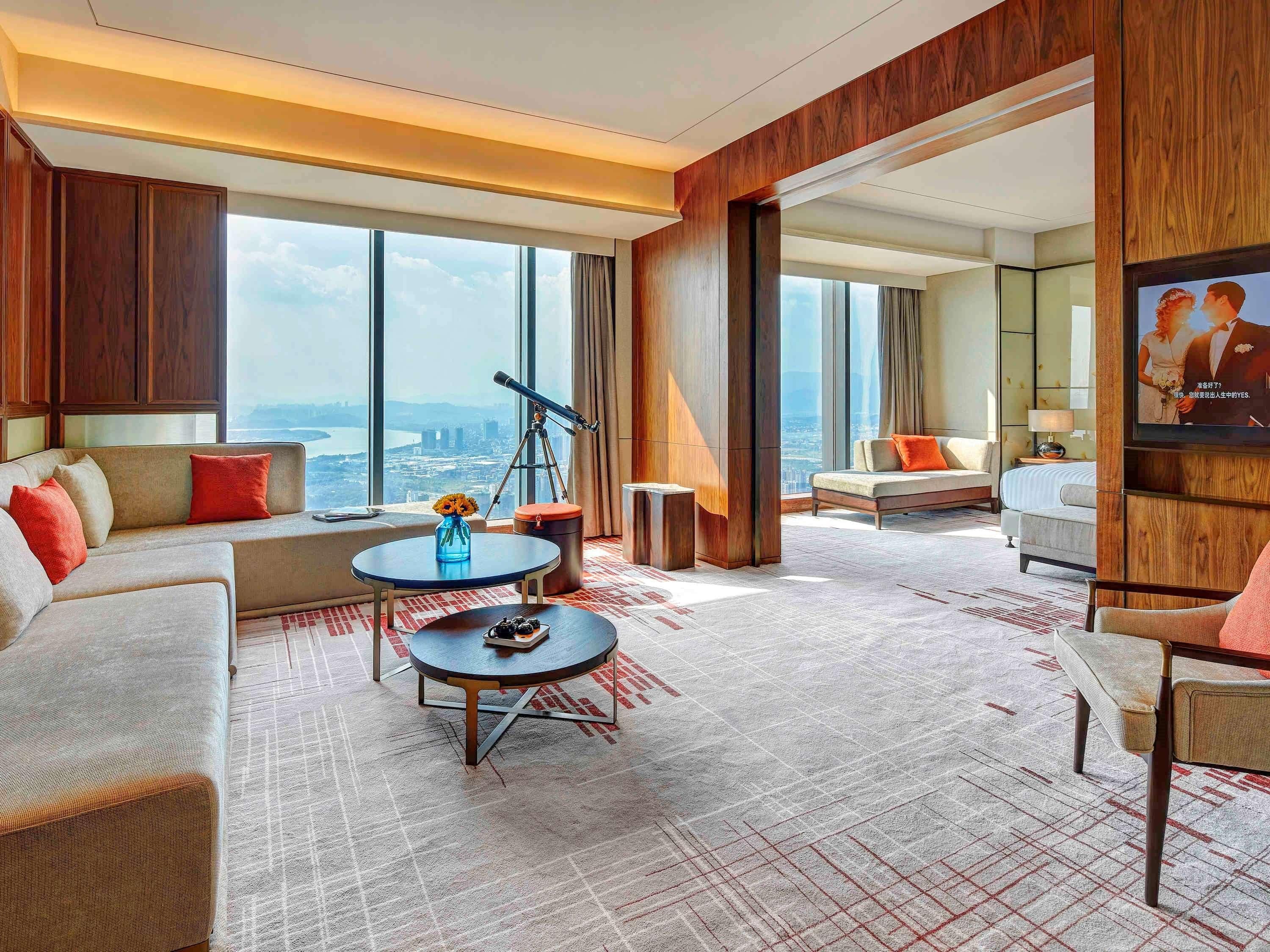 A modern hotel room with a large window, a telescope, a sofa, a coffee table, and a TV.