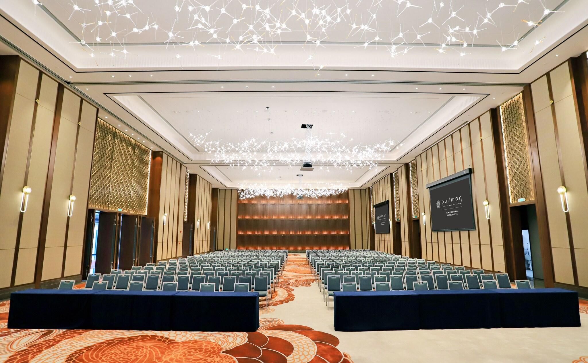 Ein Konferenzraum mit Stuhlreihen, einer Bühne und einer Decke mit hängenden Lampen.