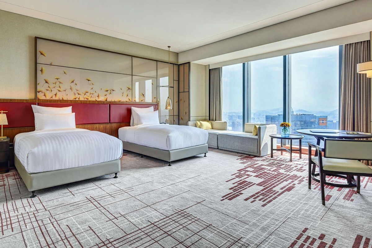 A hotel room with two beds, a sofa, a dining table, and a large window offering a city view.