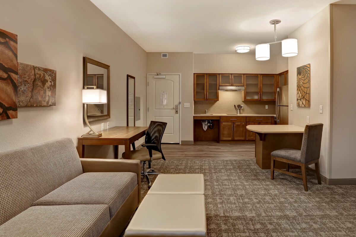 A hotel room with a sofa, a desk, a chair, a kitchenette, and artwork on the walls.