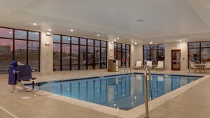 A large indoor swimming pool with clear glass walls, a blue pool, and a chair with a towel. - Staybridge Suites Overland Park - Kansas City S (Overland Park)
