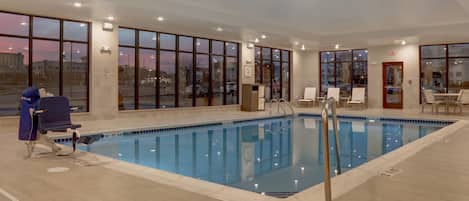 A large indoor swimming pool with clear glass walls, a blue pool, and a chair with a towel.
