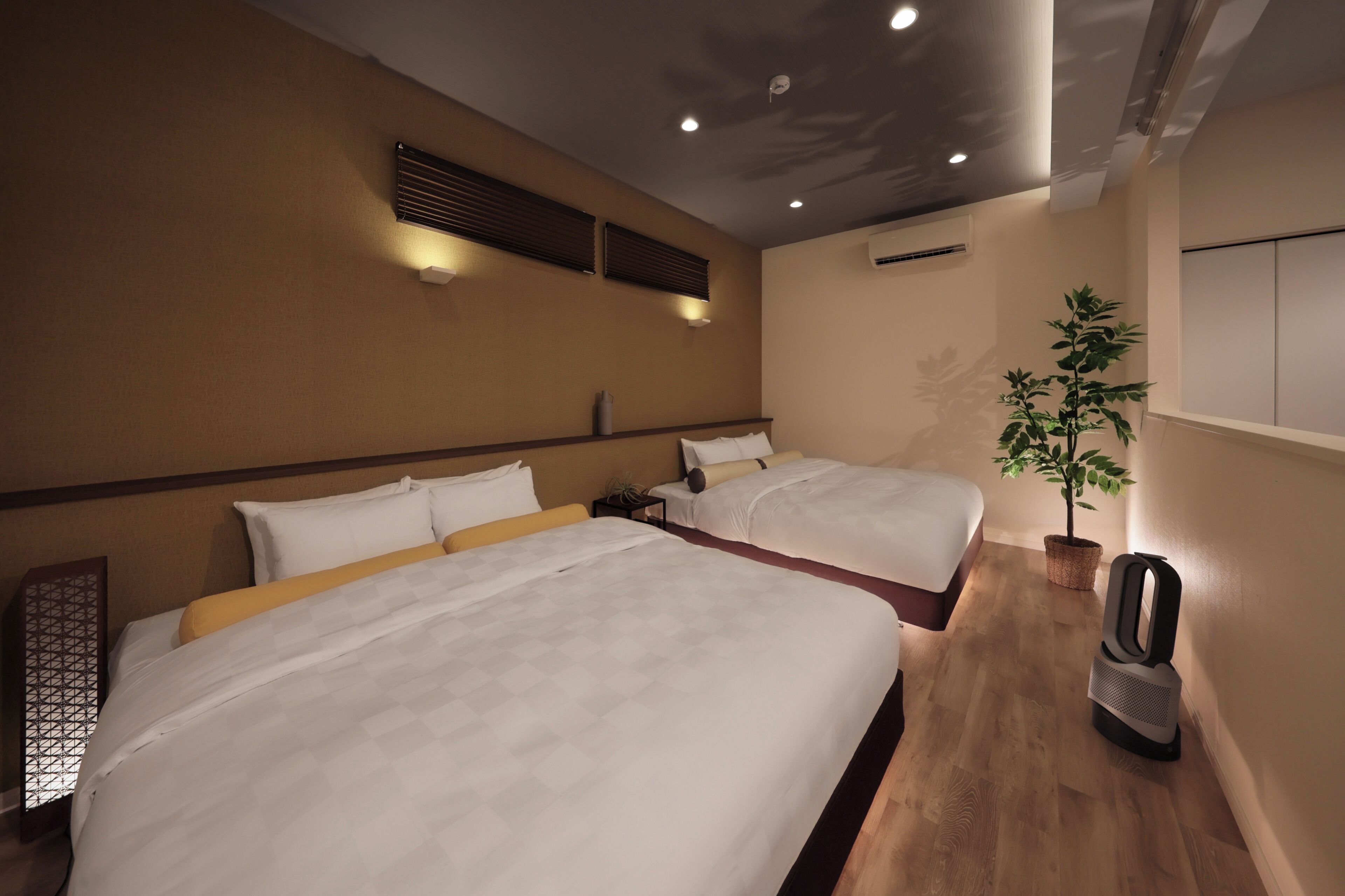 A hotel room with two beds, a wooden floor, a potted plant, a nightstand, and a ceiling fan.