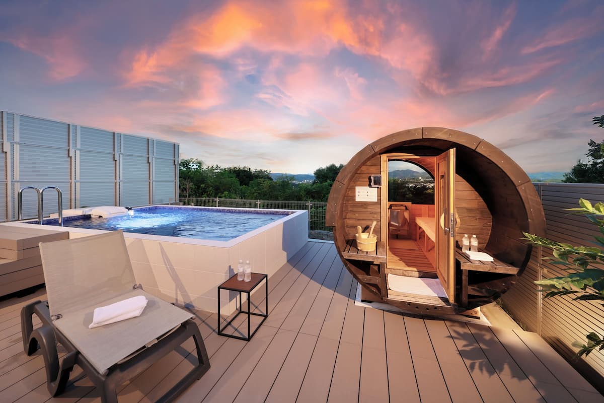 A wooden hot tub on a deck with a lounge chair and a small table, surrounded by a wooden structure with a bench.