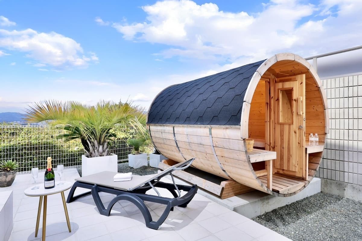 A wooden barrel-shaped sauna with a bench and a table with glasses and a bottle.