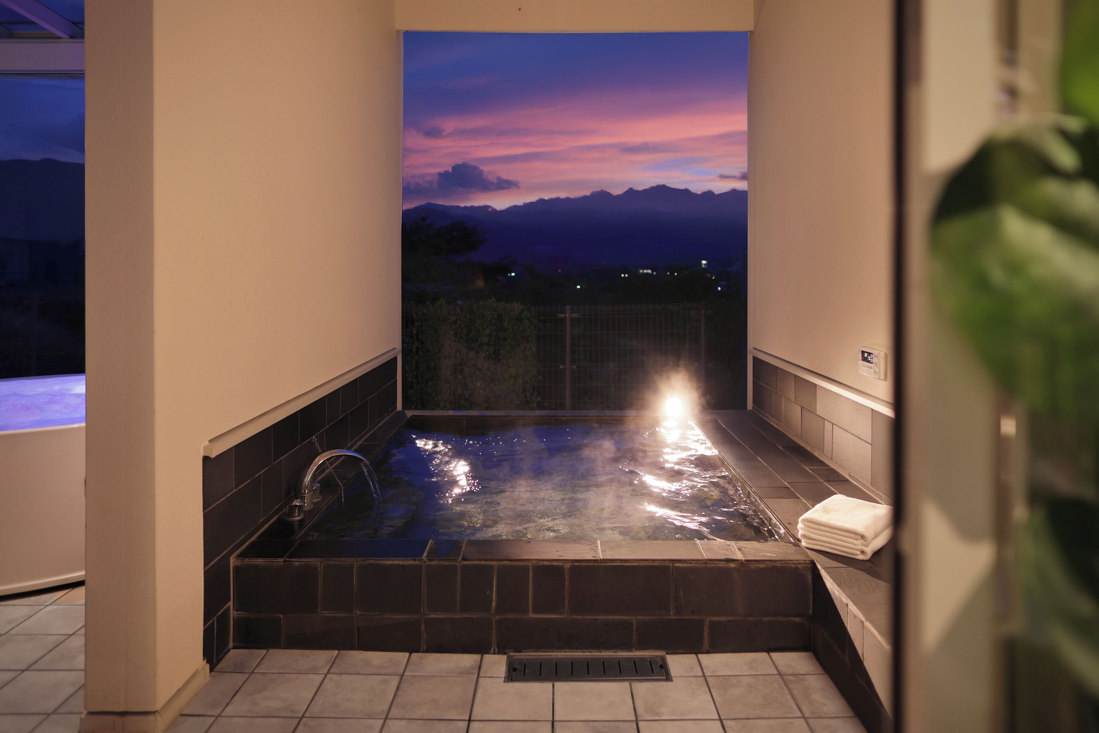 A hot tub with a view of mountains at sunset, tiled walls, and a towel on the side.