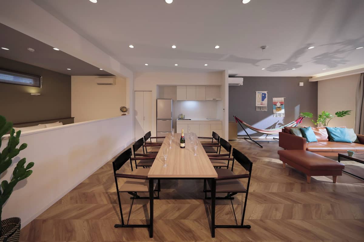 A modern dining area with a long wooden table, multiple chairs, and a kitchen in the background.