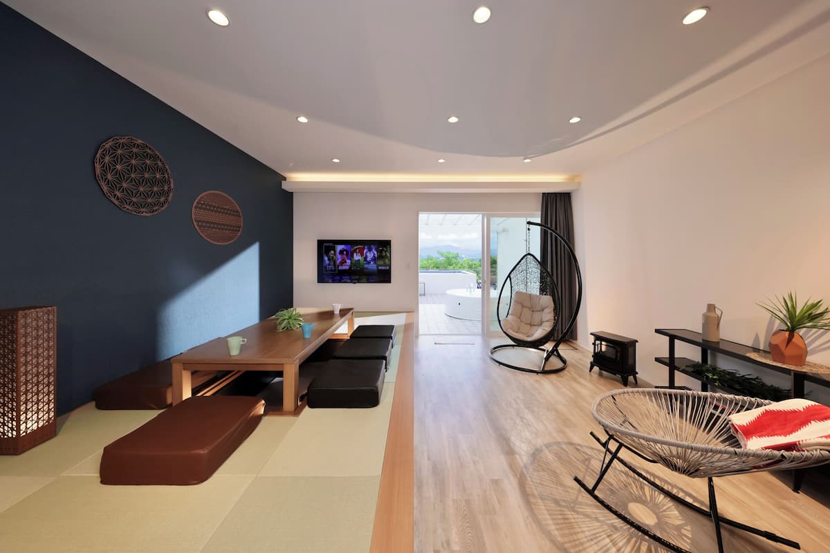 A modern living room with a wooden table, black ottomans, a hanging chair, and a television on the wall.