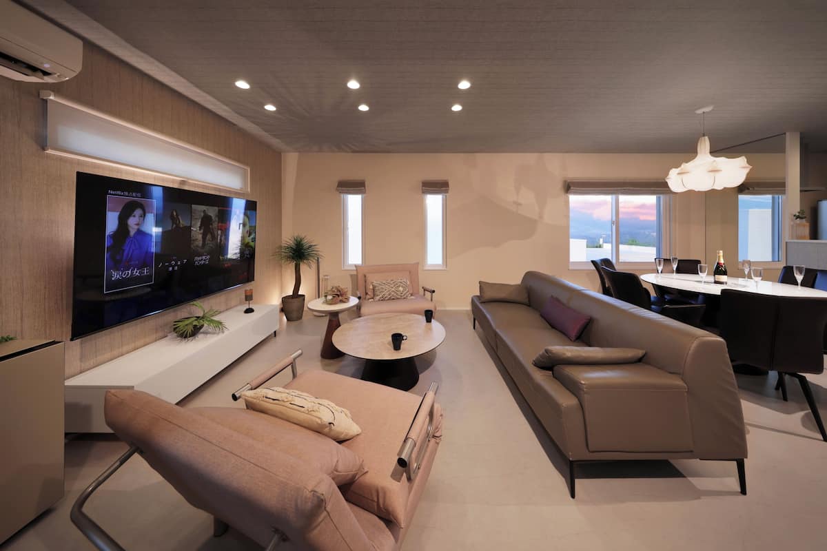 A modern living room with a flat-screen TV, a sofa, a coffee table, and a dining area with a chandelier.