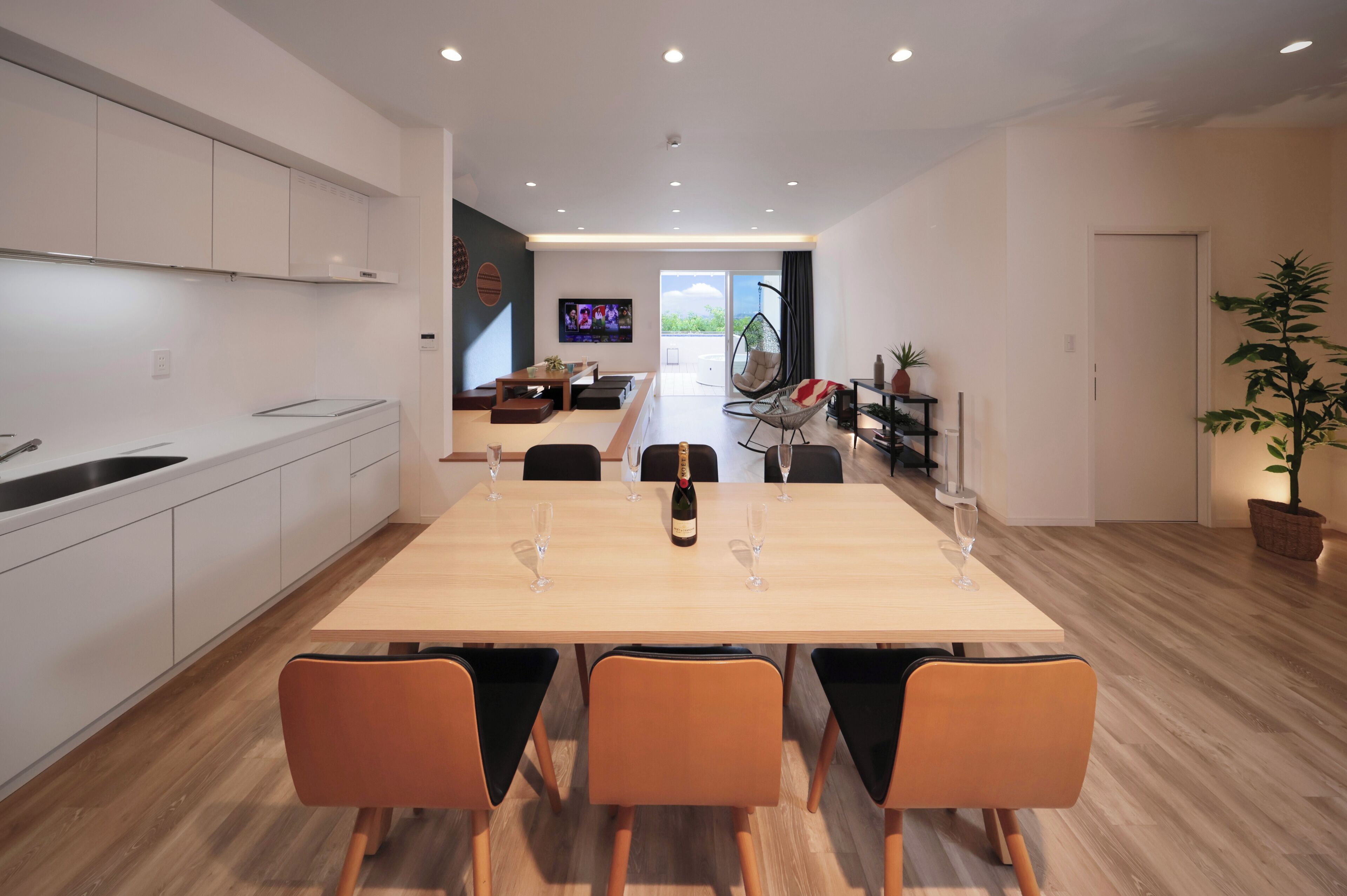 Une cuisine moderne avec des meubles blancs, un coin repas avec une table en bois et des chaises orange, un téléviseur en arrière-plan et une grande plante en pot.