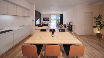 Une cuisine moderne avec des meubles blancs, un coin repas avec une table en bois et des chaises orange, un téléviseur en arrière-plan et une grande plante en pot.