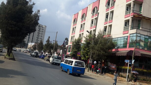 A street with a blue and white van parked on the side, buildings with balconies, and pedestrians. - SGS Hotel (Addis Ababa)