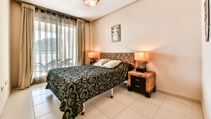 Ein Schlafzimmer mit Bett, zwei Nachttischlampen, einem Kopfteil, einem Fenster mit Vorhängen und einem Wandbild.