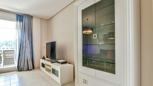 Ein modernes Wohnzimmer mit einem Flachbildfernseher auf einer weißen Unterhaltungsanlage, einem gläsernen Schrank mit Regalen und einem großen Fenster mit Vorhängen.