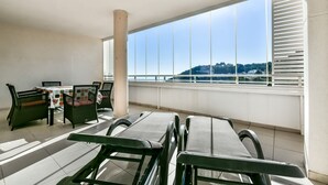 Ein Balkon mit Tisch und Stühlen, große Fenster mit Jalousien und Blick auf eine Hügel- und Stadtlandschaft.