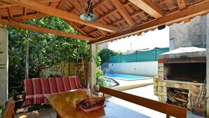 Une terrasse en bois avec un banc et une table, un barbecue en pierre et, au fond, une piscine.