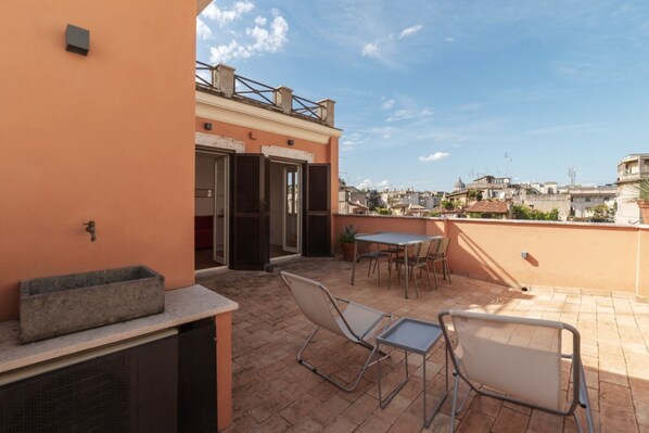 A rooftop terrace with a table and chairs, a grill, and a view of the cityscape.