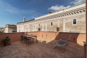A rooftop terrace with a table and chairs, potted plants, and a view of a historic building with arched windows and a chimney.