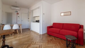 A modern living room with a red sofa, wooden herringbone flooring, a dining area with a wooden table and white chairs, and a kitchen area with white cabinets. - Modern studio with panoramic terrace, by Campo de’ Fiori - Wifi, Independent Heating, A/C (Roma)