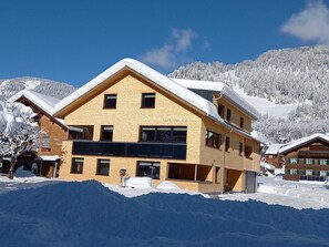 A two-story wooden building with a sloped roof, labeled 'Feriwohnung,' surrounded by snow-covered mountains. - Haus Bianca - Apartment 2-8 pers. (Schoppernau)