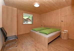 Une pièce aux murs lambrés, comprenant un lit, une chaise et une fenêtre donnant sur une maison.