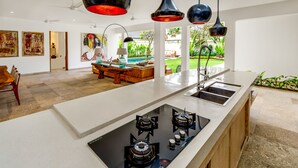 A modern kitchen with a central island, a dining area with a wooden table and chairs, and a view of a garden. - Villa Momentai (Canggu)