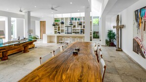 A spacious dining area with a long wooden table and white chairs, a kitchen in the background, and a large window providing natural light. - Villa Momentai (Canggu)